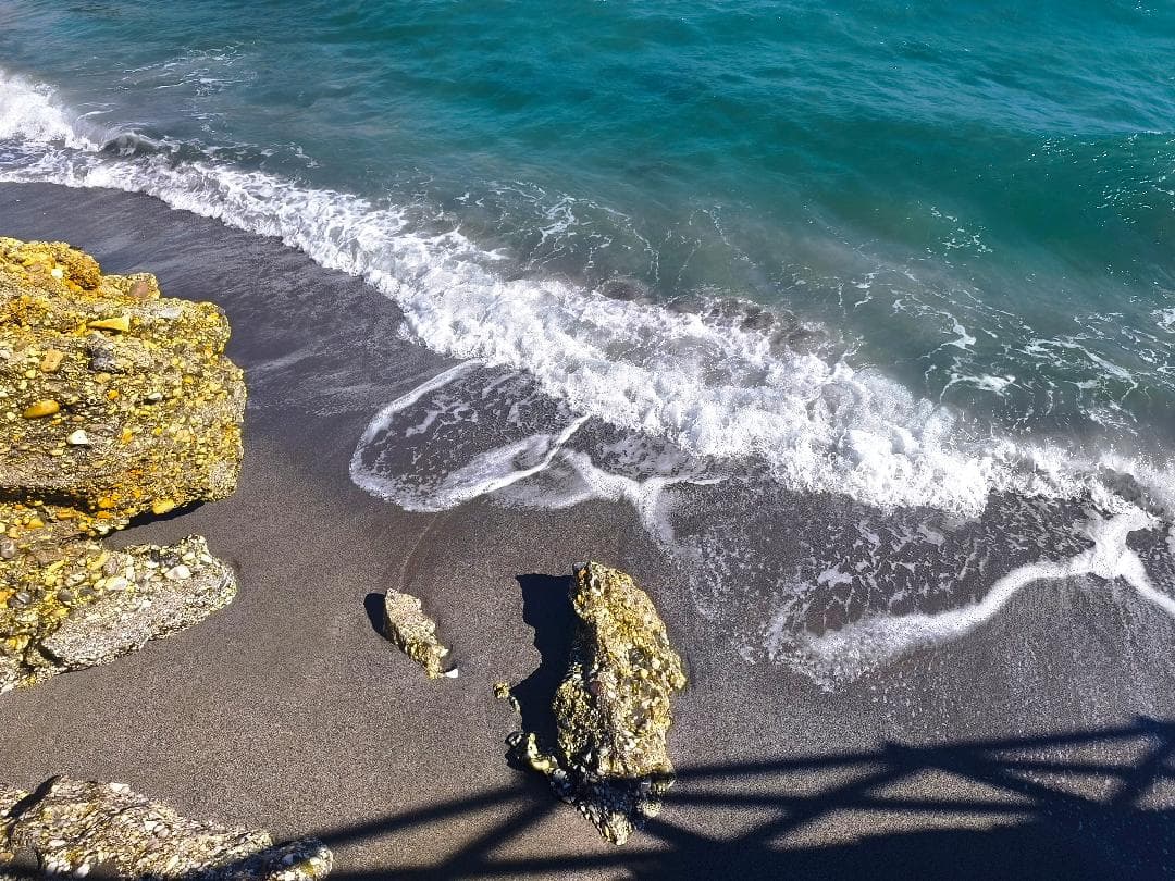 Olas de mar turquesa rompiendo sobre arena oscura y rocas amarillas en la orilla.