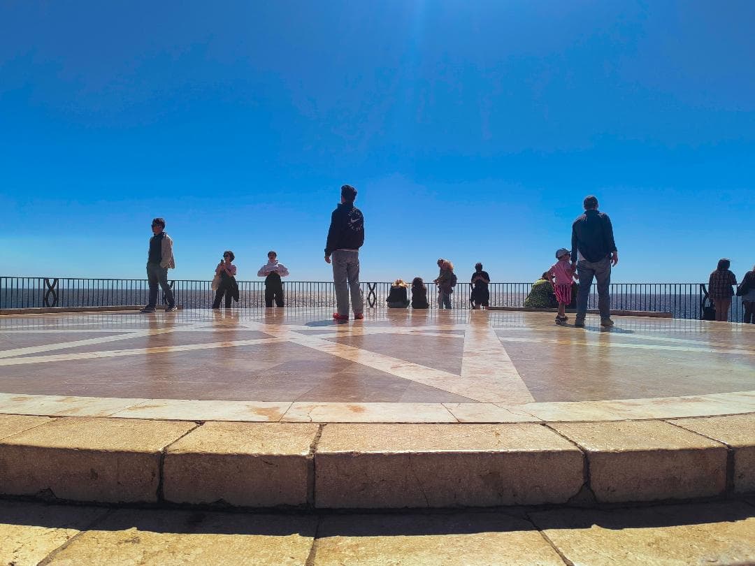 Personas en un mirador de piedra frente al mar bajo un cielo azul despejado.