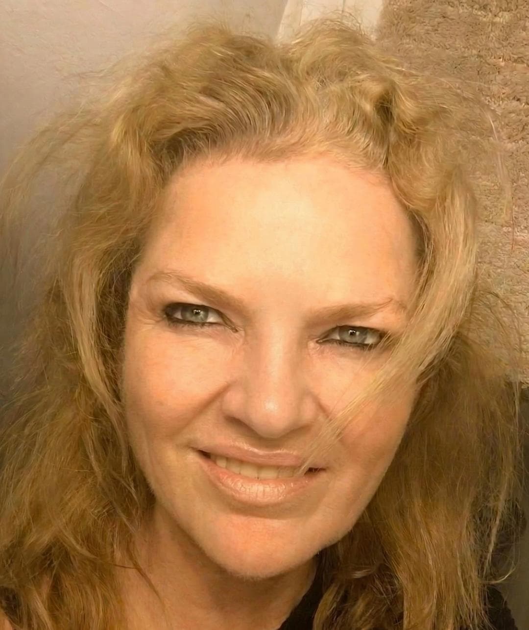 Primer plano de una mujer con cabello rubio ondulado, ojos verdes delineados y sonrisa suave.