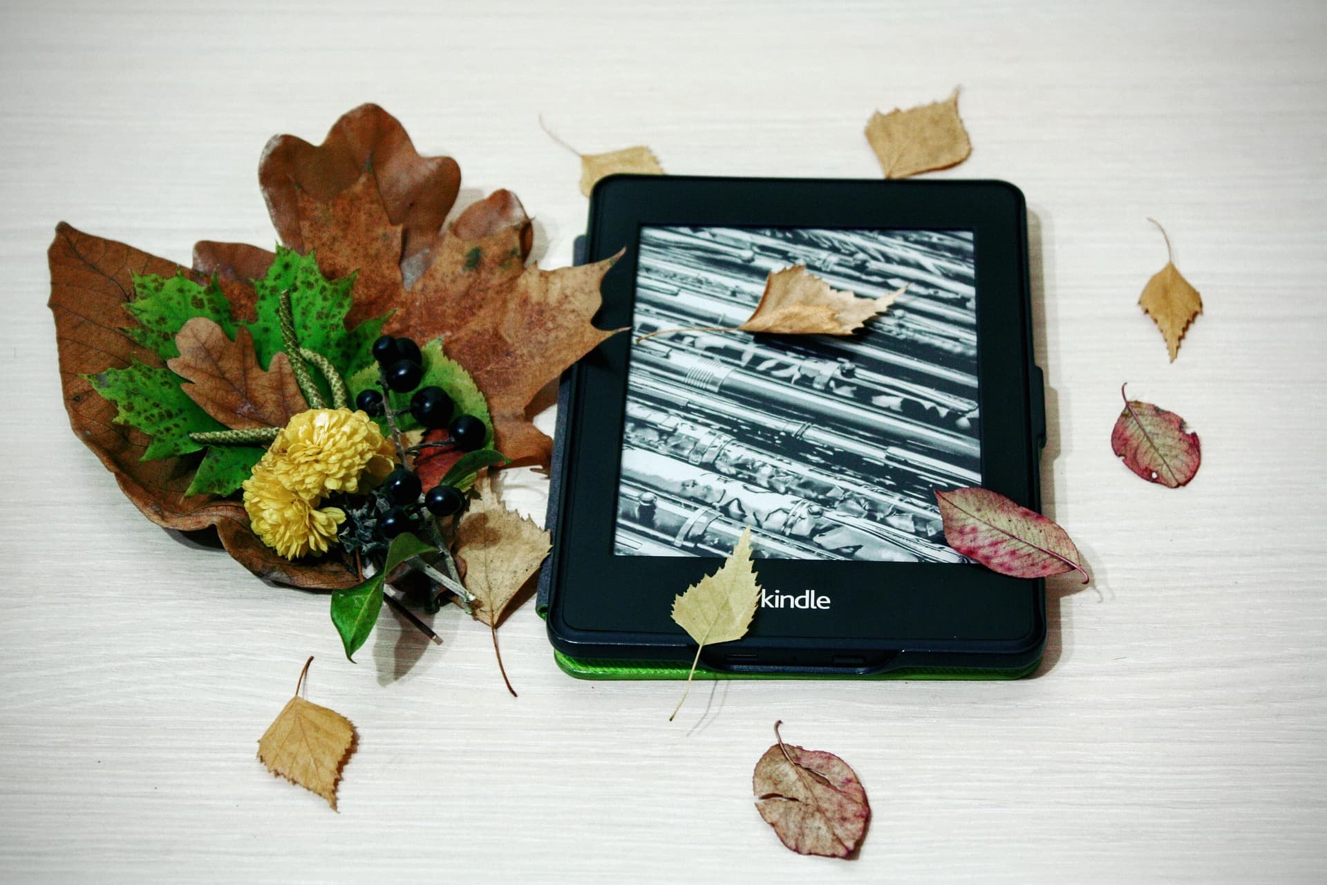 Lector Kindle rodeado de hojas secas, flores amarillas y bayas sobre una mesa clara.