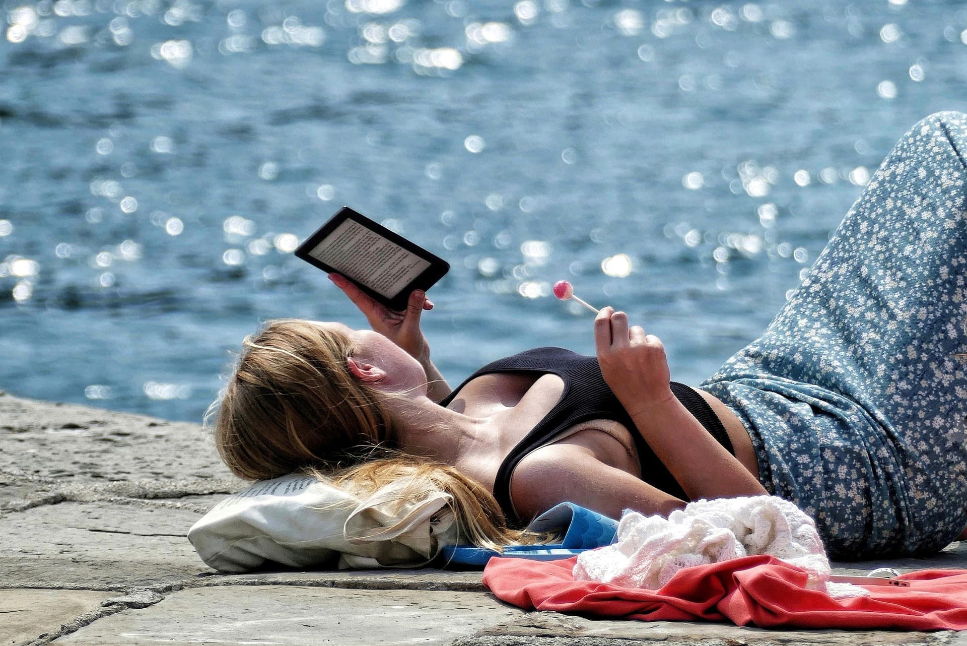 Mujer tumbada frente al mar leyendo un libro electrónico y sosteniendo una piruleta rosa.